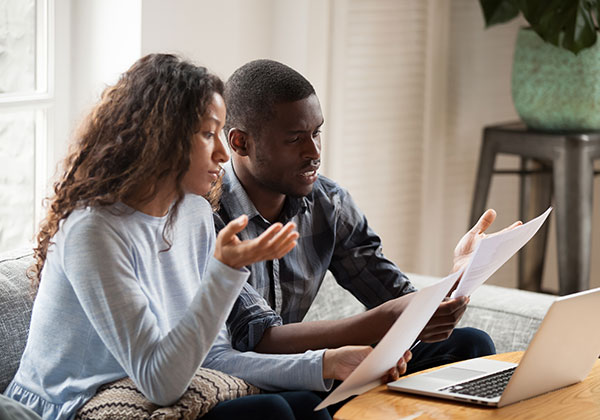 Young mixed-race couple holding papers, comparing notes, on a couch at a table with a laptop computer.